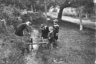 Kinder in Matrosenanzug und Matrosenkleid beim Spiel im Freien mit einem Wasserrad. 