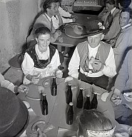 Ein Dorffest: Einige Männer und Frauen, in Tracht gekleidet, sitzen an einem runden Tisch. Auf dem Tisch stehen Flaschen und Speisen 