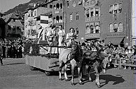 Ein folkloristischer Umzug: Auf einem geschmückten, von Pferden gezogenen Umzugswagen stehen drei Frauen in traditioneller Kleidung und eine Art Weltkugel, auf der das Bozner Stadtwappen aufgesetzt ist. 